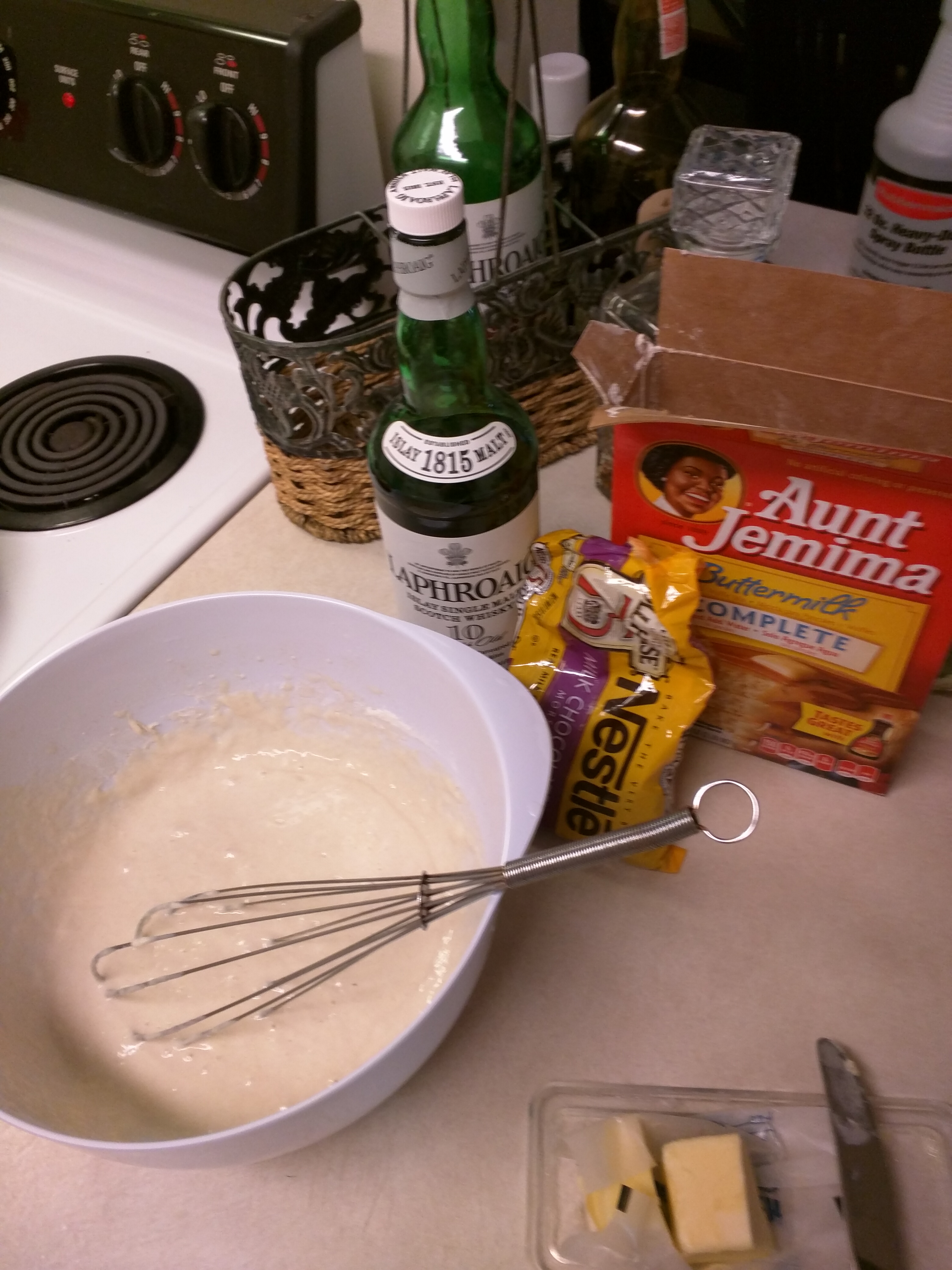 Pankake mix, batter, whisk, chocolate chips, and several bottles of whiskey next to a stovetop.